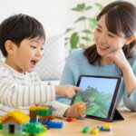 ブロック遊びやゲーム画面を通して英語にふれる幼児と母親の明るい家庭の場面