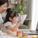 幼児が自宅で母親に見守られながらオンラインで英語の先生に手を振る場面