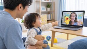 父親のそばで画面の先生の人形を見つめる幼児のイメージ（AI生成）