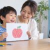 母親と子どもが描いたリンゴの絵を画面に見せるイメージ（AI生成）