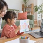 母親の隣でオンラインの先生に絵を見せる子どものイメージ（AI生成）
