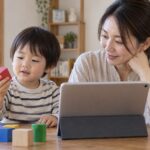 母親の隣で画面ではなく手元の積み木を見る子どものイメージ（AI生成）