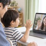 父親と一緒にオンラインの先生へ手を振る幼児のイメージ（AI生成）