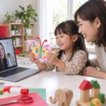 母親の隣で工作を見せ、画面越しの先生とやりとりする幼児のイメージ
