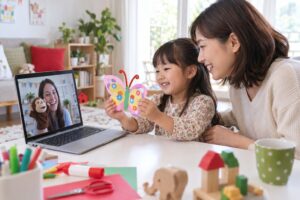 母親の隣で工作を見せ、画面越しの先生とやりとりする幼児のイメージ
