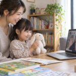 母親の隣でぬいぐるみを持ち、画面の先生に微笑む幼児のイメージ