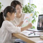 母親が見守る家の机で、海外の友だちに絵を見せる小学生のイメージ