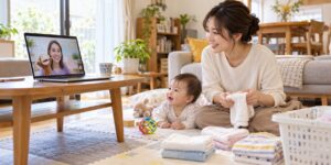 赤ちゃんのすぐ隣でタオルをたたみながらオンラインの先生を見る母親のイメージ（AI生成）