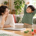 閉じたパソコンから離れて母親の隣で伸びをする子どものイメージ（AI生成）