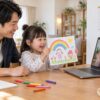 父親の隣でオンラインの先生に描いた絵を見せる幼児のイメージ（AI生成）